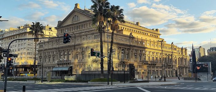 The Teatro Colon seen across an empty street corner at low sun, its ochre stone facade and tiers of arched windows lit gold, tall palms rising in front of it, traffic lights and a wide zebra crossing in the foreground under a blue sky with scattered cloud.
