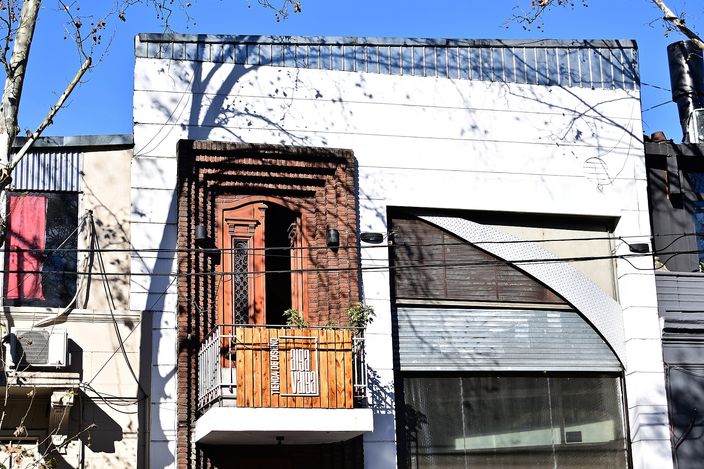 A narrow two-storey house in Palermo with a tall arched wooden door and a small first-floor balcony, framed by bare tree branches against a white facade under blue sky