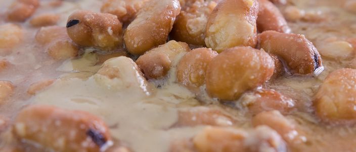 Extreme close-up of ful medames: whole and split brown fava beans in a thick, glossy broth, skins burst on many, a sheen of oil across the surface.