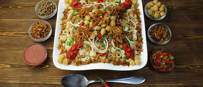A square white plate heaped with koshari — layered rice, brown lentils and short macaroni under tomato sauce and a heap of deep-fried onions — ringed by small glass bowls of chickpeas, extra fried onions, a garlic-vinegar dakka sauce and a red chili shatta sauce, with whole chilies and garlic cloves and a spoon laid in front, on a dark wood table.