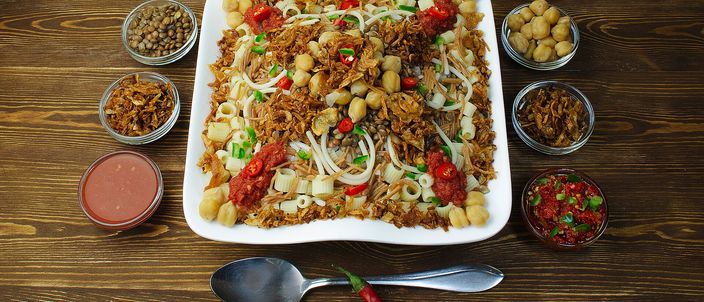 A square white plate heaped with koshari — layered rice, brown lentils and short macaroni under tomato sauce and a heap of deep-fried onions — ringed by small glass bowls of chickpeas, extra fried onions, a garlic-vinegar dakka sauce and a red chilli shatta sauce, with whole chillies and garlic cloves and a spoon laid in front, on a dark wood table.