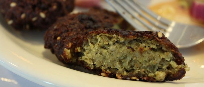 A fava-bean falafel broken open on a white plate, its interior bright green against the dark sesame-studded crust, with two whole patties and a fork behind.