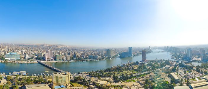 Wide aerial panorama of central Cairo at midday, the Nile River crossed by bridges with dense high-rise skyline on both banks and a slim tower-block hotel in the foreground.