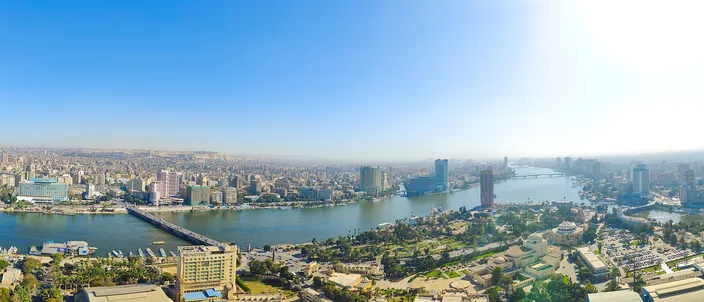 Wide aerial panorama of central Cairo at midday, the Nile River crossed by bridges with dense high-rise skyline on both banks and a slim tower-block hotel in the foreground.