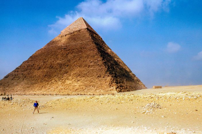 The Pyramid of Khafre seen from ground level on a clear day, a lone visitor walking across the sand in front of it and a band of smooth original casing stone still capping its summit.