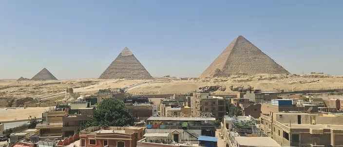 A panoramic view from the edge of Giza town: three pyramids of the Giza plateau rising directly behind a dense row of apartment rooftops, with no open desert buffer between the two.