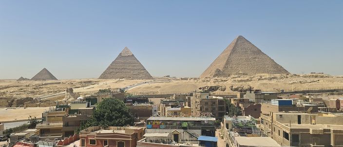 A panoramic view from the edge of Giza town: three pyramids of the Giza plateau rising directly behind a dense row of apartment rooftops, with no open desert buffer between the two.