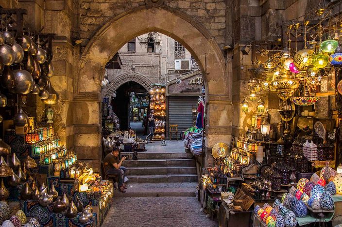 Inside the covered lanes of Khan el-Khalili market: brass lanterns and lamps hanging from stalls on both sides of a stone archway, with shoppers under strings of bunting further down the passage.