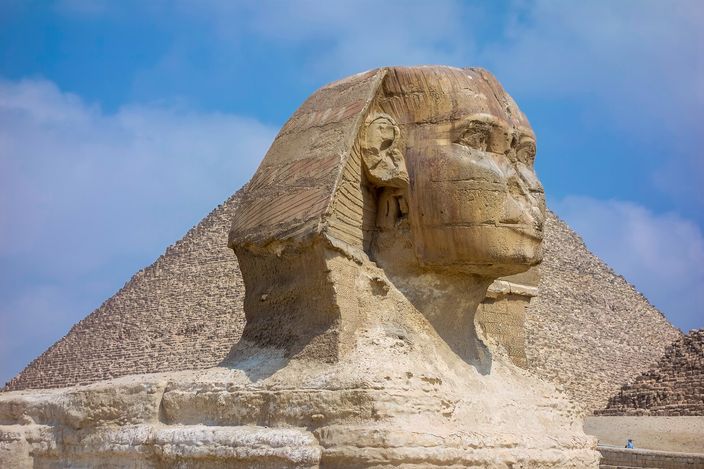 The Great Sphinx of Giza in close profile, its face and paws in the foreground with one of the Giza pyramids rising behind it against a clear blue sky.
