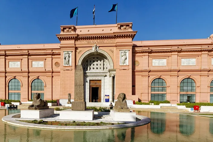 The pink neoclassical facade of the Egyptian Museum on Tahrir Square, its reflecting pool and flanking sphinx statues in front of the entrance under a clear sky.