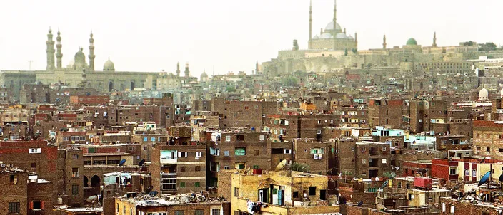 A dense rooftop panorama of old Cairo, with the domes and twin minarets of Islamic-Cairo mosques and a distant domed silhouette rising above tightly packed apartment buildings.