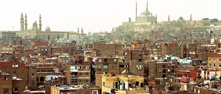 A dense rooftop panorama of old Cairo, with the domes and twin minarets of Islamic-Cairo mosques and a distant domed silhouette rising above tightly packed apartment buildings.