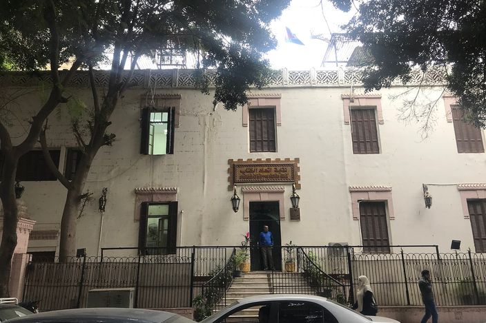 A white colonial-era building with iron balconies on a tree-shaded Zamalek street, cars parked along the kerb and two pedestrians passing.