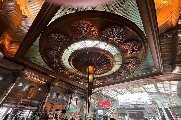 The ornate fan-patterned domed ceiling of Cairo's Ramses railway station concourse, with escalators, passengers and station signage below.
