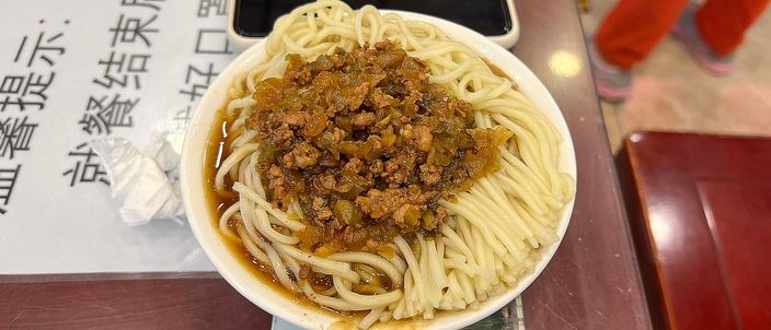 A bowl of dry dan dan noodles: thin wheat noodles topped with a mound of dark, crisp minced pork and preserved mustard stem, barely any liquid in the bowl.
