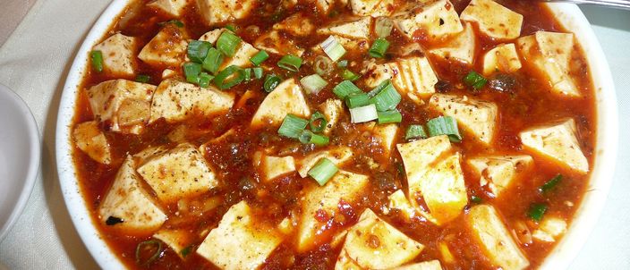 A plate of mapo tofu: white cubes of soft tofu in a glossy red-brown chilli sauce, scattered with chopped green spring onion.