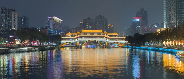 The illuminated Anshun Bridge, a traditional covered-arch pavilion glowing gold, spanning the Jin River at night with colorful reflections shimmering on the water and the city skyline behind.