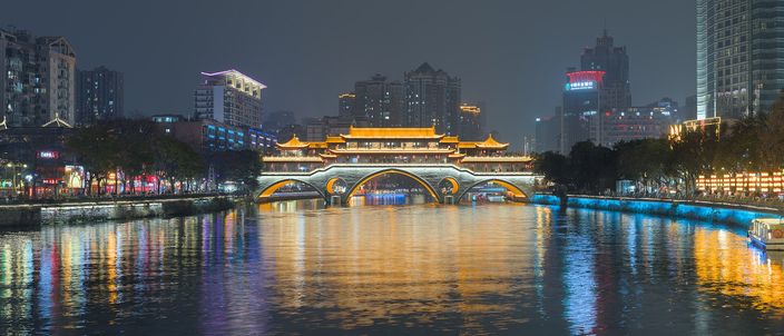 The illuminated Anshun Bridge, a traditional covered-arch pavilion glowing gold, spanning the Jin River at night with colourful reflections shimmering on the water and the city skyline behind.