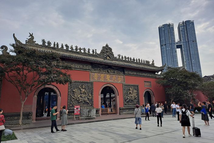 The red-walled entrance gate of Daci Temple, with a carved frieze and a tiled, upswept traditional roof, people crossing the paved forecourt in front of it, and two tall modern glass towers rising directly behind.