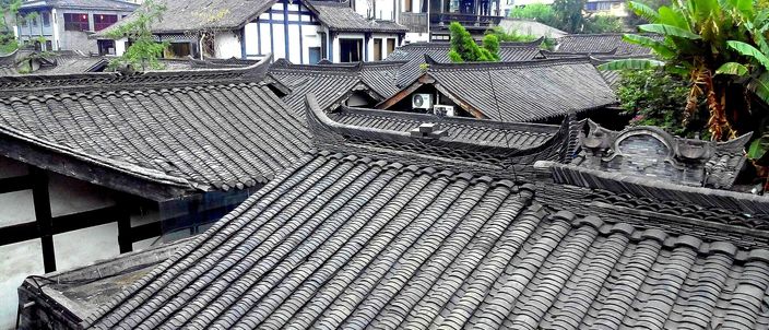 Grey barrel-tiled rooftops of low restored courtyard houses seen from above, with a white-walled dark-timbered two-storey building, leafy trees and a palm rising behind them.