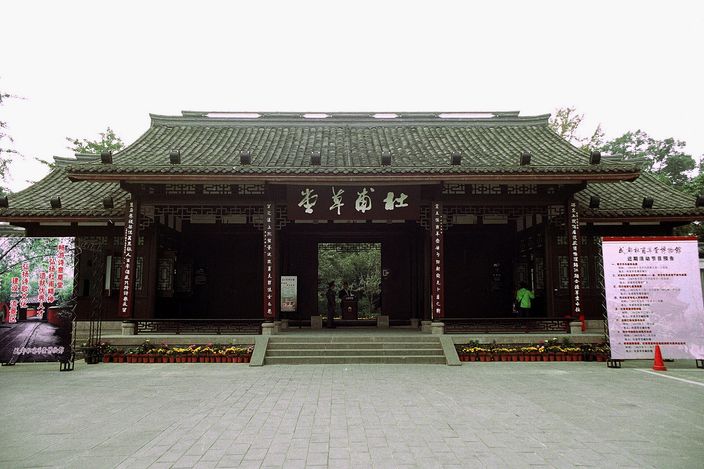 The wide timber-and-tile entrance hall of a walled garden, its grey double-eaved roof over dark lacquered doors and a horizontal plaque of gold Chinese characters, with flower beds and stone steps across an empty paved forecourt.