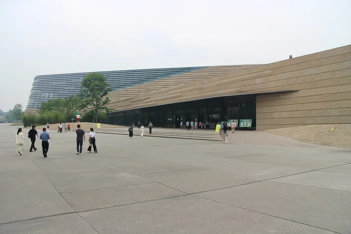 A long low museum building with a steeply sloping wall of horizontal sand-colored bands and a dark glazed roof folding down to the ground, seen across a wide empty concrete plaza with a scatter of visitors walking towards its entrance.