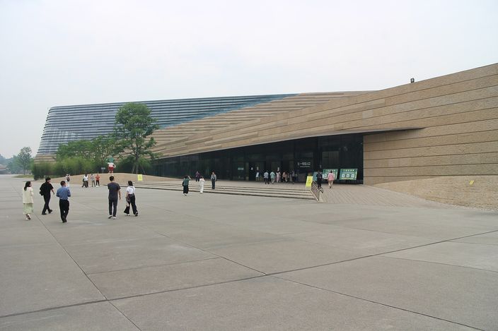 A long low museum building with a steeply sloping wall of horizontal sand-coloured bands and a dark glazed roof folding down to the ground, seen across a wide empty concrete plaza with a scatter of visitors walking towards its entrance.