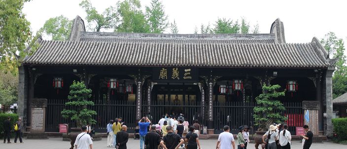 A long single-storey temple hall with a sweeping grey tiled roof, dark timber columns and a red-charactered plaque over its central doorway, seen across a paved forecourt with visitors walking towards it and trees rising behind.