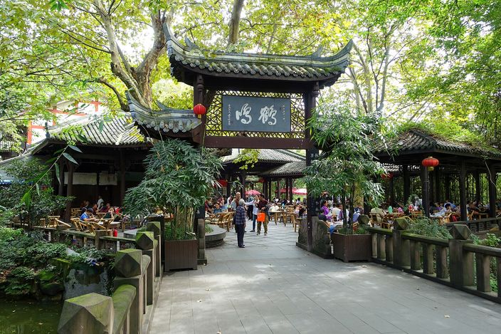 An open-air teahouse in People's Park, Chengdu: rows of bamboo chairs and low tables under leafy trees, with a dark carved wooden pavilion gateway hung with red lanterns and Chinese characters, people strolling a stone path in the foreground.