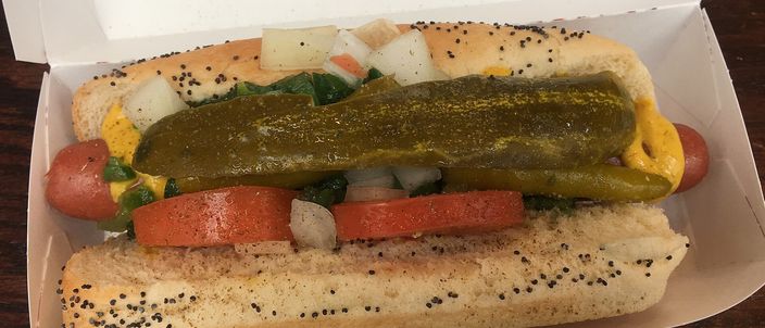 A Chicago-style hot dog in a poppy-seed bun, dressed with yellow mustard, green relish, chopped onion, tomato wedges, a dill pickle spear and sport peppers, served in an open paper box.