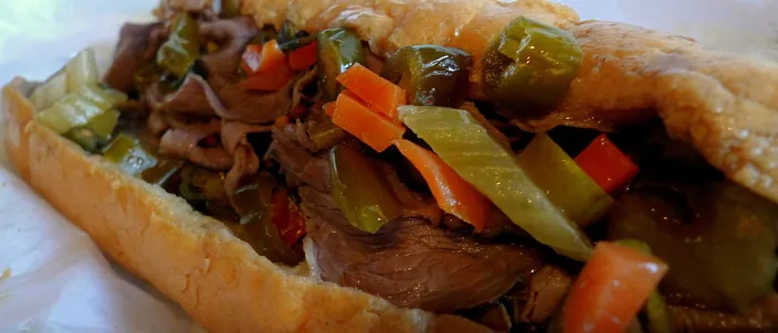 A Chicago Italian beef sandwich on a long roll, piled with thin-sliced beef and heaped with colorful hot giardiniera of pickled peppers, celery and carrot.