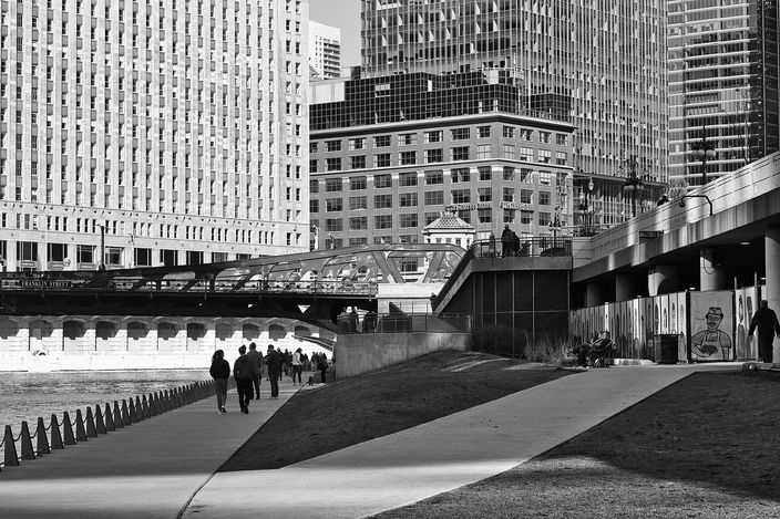 Black-and-white view along the Chicago Riverwalk: people walking a paved riverside path a level below the street, a bridge carrying an elevated train across the middle of the frame, and Loop office towers rising behind.