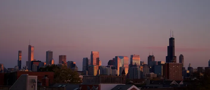The Chicago skyline at dusk seen over the rooftops and chimneys of a low-rise neighborhood, the Willis Tower's dark twin-antenna profile standing at the right against a pink and violet sky.