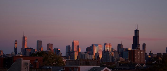 The Chicago skyline at dusk seen over the rooftops and chimneys of a low-rise neighbourhood, the Willis Tower's dark twin-antenna profile standing at the right against a pink and violet sky.