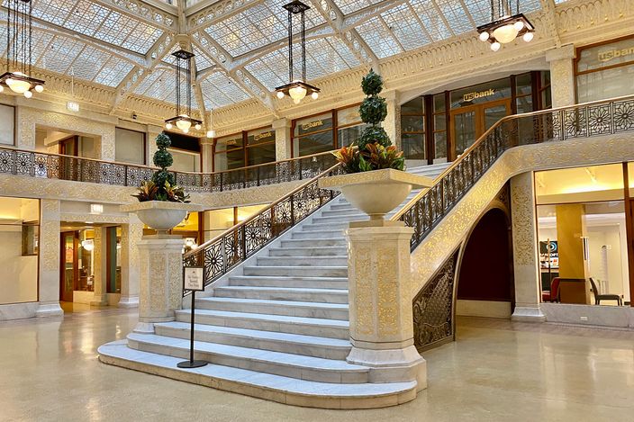 The two-storey light court of the Rookery Building: a glazed roof of geometric white ironwork over a broad white marble staircase, pierced gilt balustrades running round the mezzanine, and planted urns on piers at the foot of the stairs.