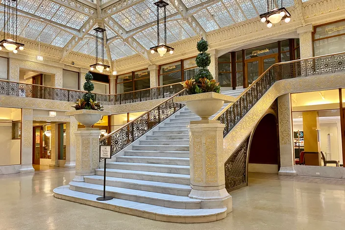 The two-story light court of the Rookery Building: a glazed roof of geometric white ironwork over a broad white marble staircase, pierced gilt balustrades running round the mezzanine, and planted urns on piers at the foot of the stairs.