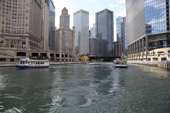 The Chicago River from water level on a bright day, two small white sightseeing boats crossing the channel, stone and glass towers rising on both banks, a bridge and the setback crown of the Jewelers Building beyond.