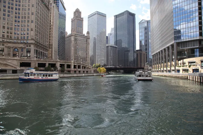 The Chicago River from water level on a bright day, two small white sightseeing boats crossing the channel, stone and glass towers rising on both banks, a bridge and the setback crown of the Jewelers Building beyond.