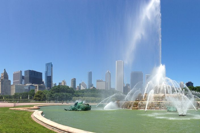 Buckingham Fountain in Grant Park under a clear sky, its central jet thrown high above the tiered basins and the bronze seahorses, with the Loop's towers ranged along the horizon behind and the curved pink walkway in the foreground.