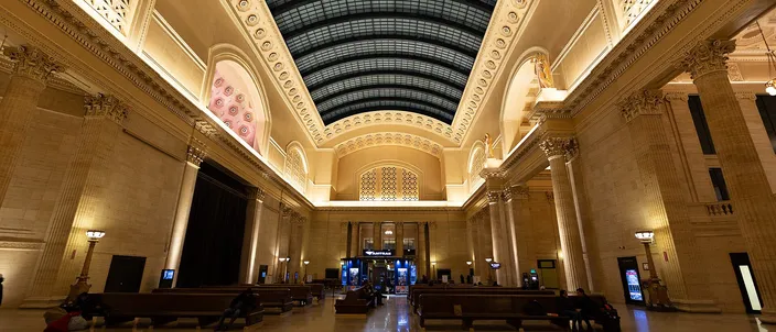 The vaulted Great Hall of Chicago's Union Station: a long barrel skylight of dark glazed panels above a cream stone hall, tall Corinthian columns down both sides, and wooden benches on a polished marble floor with a few travelers waiting on them.