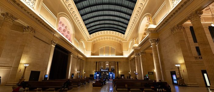 The vaulted Great Hall of Chicago's Union Station: a long barrel skylight of dark glazed panels above a cream stone hall, tall Corinthian columns down both sides, and wooden benches on a polished marble floor with a few travellers waiting on them.