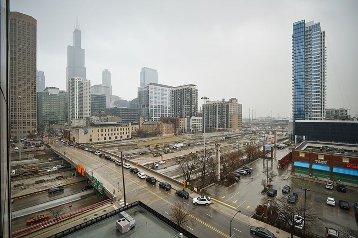 Overcast daytime view from a hotel window over the West Loop: wide multi-lane avenues and a rail corridor below, low brick buildings, and the Willis Tower and downtown high-rises rising through grey haze behind.