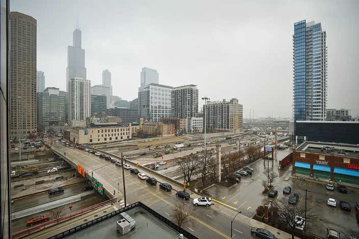 Overcast daytime view from a hotel window over the West Loop: wide multi-lane avenues and a rail corridor below, low brick buildings, and the Willis Tower and downtown high-rises rising through gray haze behind.