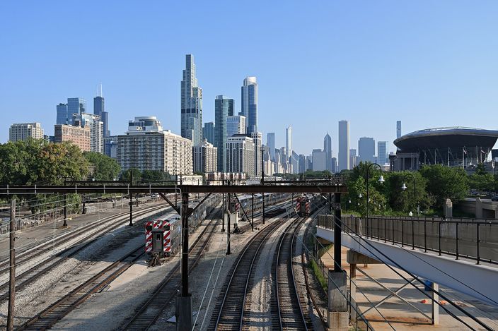Multiple commuter rail tracks curving toward the Chicago skyline in morning light, seen from a pedestrian bridge, with a stadium and clustered downtown towers on the horizon.