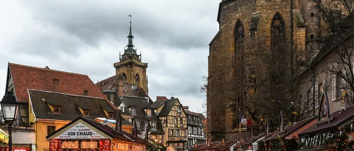 Christmas market stalls under strings of lights on place des Dominicains in Colmar, half-timbered houses behind and the green-roofed tower of the Collégiale Saint-Martin rising above them