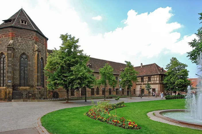 The Gothic chapel and long convent buildings of the Unterlinden Museum in Colmar beyond a lawn, flower beds and a fountain