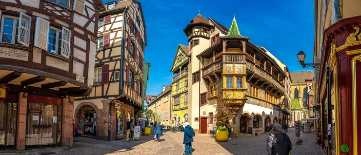 Wide view of the Maison Pfister at the corner of Rue des Marchands in Colmar, its painted facade, wooden gallery and corner turret under a deep blue sky