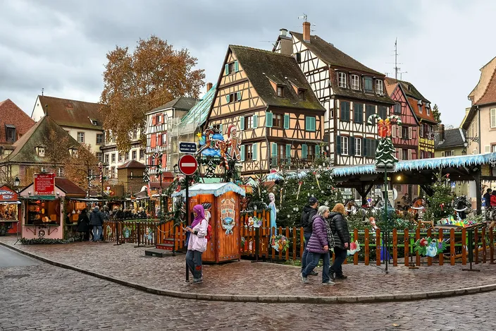 Half-timbered houses around a cobbled square in Colmar's Little Venice, the children's Christmas market below them with its fenced nativity, small wooden huts and decorated trees