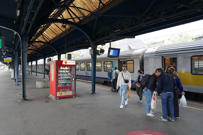 Travelers with bags on a covered platform at Colmar station beside a regional TER train bound between Strasbourg and Basel
