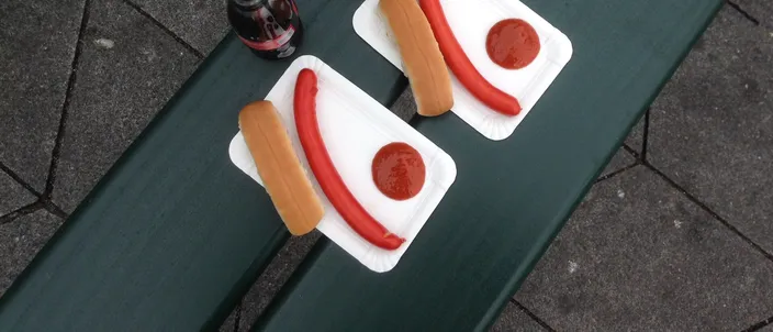 Two red sausages on paper trays, each with a soft white roll beside it and a dab of ketchup, set on a green bench next to two small bottles of chocolate milk